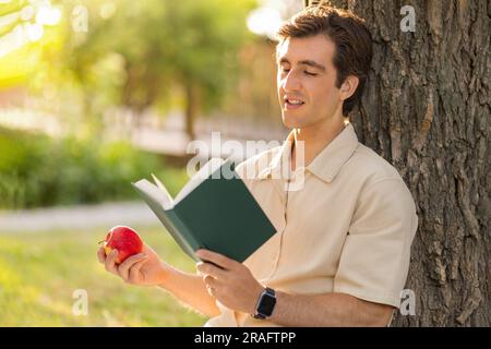 relaxed millennial man sitting under tree, reading book Stock Photo