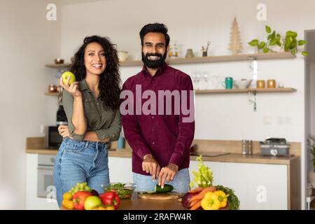 Portrait of happy arab couple posing in kitchen interior, man cooking healthy salad, woman holding apple and smiling Stock Photo