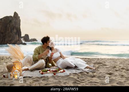 Loving young couple embracing, drinking wine and enjoying picnic date on beach, copy space Stock Photo