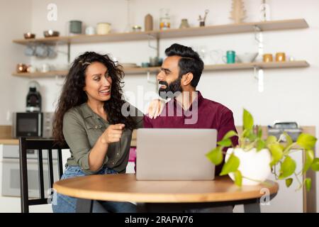 Excited middle eastern couple shopping online via laptop computer, sitting in kitchen interior, copy space Stock Photo
