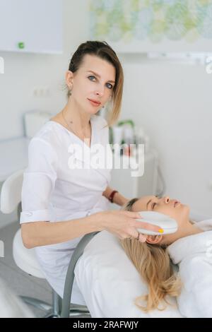 Vertical portrait of female cosmetologist doing hydro peeling procedure for woman in white bathrobe lying on medical couch in cosmetology clinic. Stock Photo