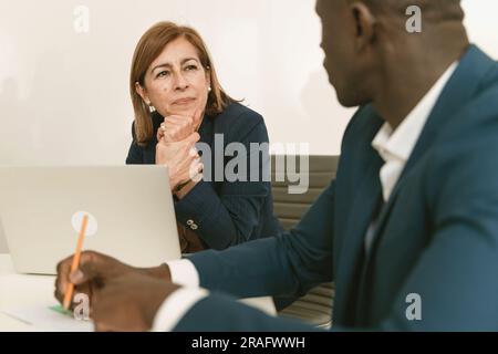 In a bright office, a senior Caucasian woman with long, straight brown hair is seen in deep concentration, hand on chin, as she listens to a young Afr Stock Photo