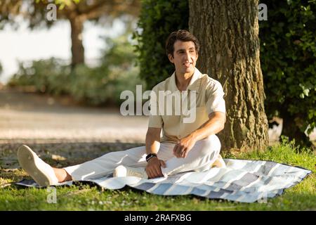 Positive handsome millennial man sitting under tree at park Stock Photo