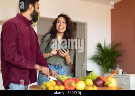 Happy arab couple cooking together, woman reading recipe on smartphone while man cutting vegetables for salad Stock Photo