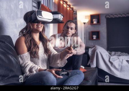 Cheerful modern couple of caucasian guys play together in living room of their studio house. Male guy enjoyng life with a girl playng videogames with Stock Photo