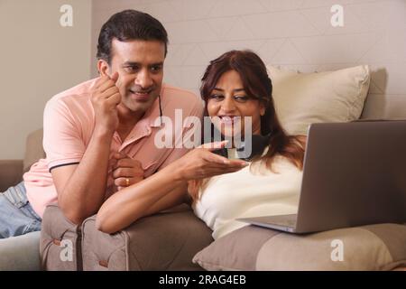 Indian happy Couple eating salad and healthy diet sitting on a sofa holding a laptop surfing online. Stock Photo