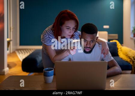 Serious family diverse couple getting ready for big deal working together with laptop in evening. Stock Photo