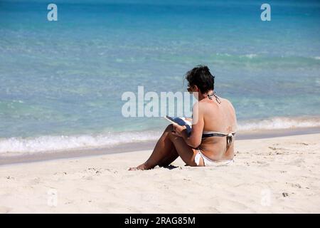 Woman in white swimsuit sitting on the sand, sunbathing and reading a paper book on sea waves background. Beach leisure on tropical coast Stock Photo