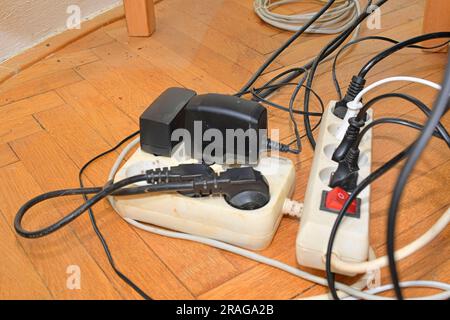 Cable chaos clutter from multiple electric wire extension cords and multi-contact plugs on wooden floor. Tangled cables on the parquet floor Stock Photo