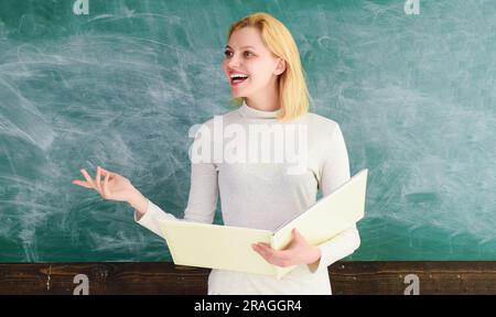 Smiling female teacher with notebook standing near blackboard giving lesson. Happy student or teacher in classroom. Education in high school Stock Photo