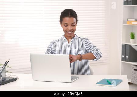 Smiling African American intern checking time on watch while working at white table in office Stock Photo