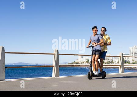 Happy biracial gay male couple riding scooter together on promenade by the sea, copy space Stock Photo