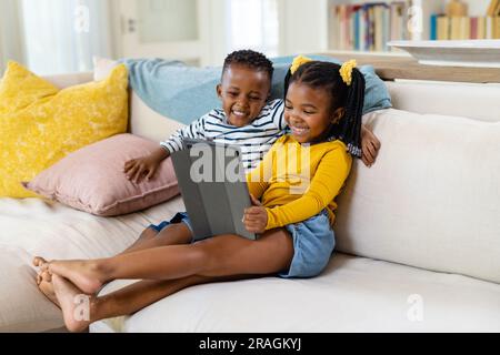 Cheerful african american siblings using digital tablet while sitting on sofa in living room Stock Photo