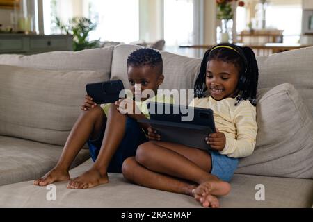African american siblings using digital tablet and smartphone while relaxing on sofa in living room Stock Photo