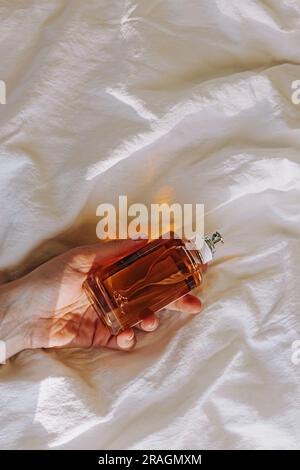 Female hand holding a bottle of perfume on the white bedding in natural light. Morning routine Stock Photo