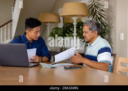 Diverse financial advisor and senior man discussing paperwork and using laptop in dining room Stock Photo