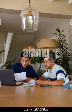 Diverse financial advisor and senior man discussing paperwork and using laptop in dining room Stock Photo