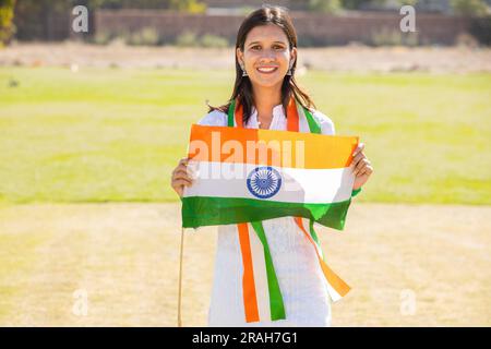 Young beautiful girl wearing traditional white dress holding indian flag while standing at park celebrating Independence day or Republic day. Stock Photo