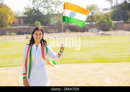 Happy Young beautiful girl wearing traditional white dress holding indian and weaving flag while standing at park celebrating Independence day or Repu Stock Photo