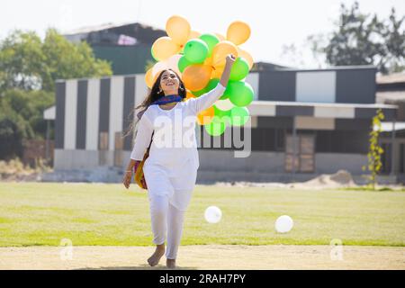 Beautiful happy young indian girl wearing traditional white kurta dress running with tricolor balloons at park celebrating independence day or republi Stock Photo