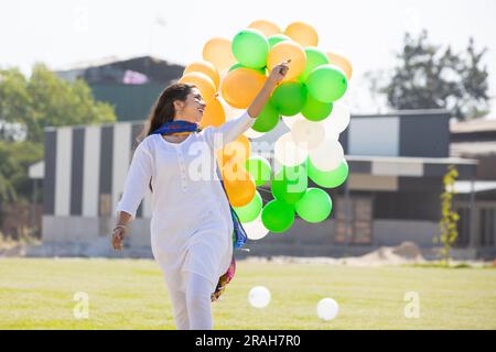 Beautiful happy young indian girl wearing traditional white kurta dress running with tricolor balloons at park celebrating independence day or republi Stock Photo