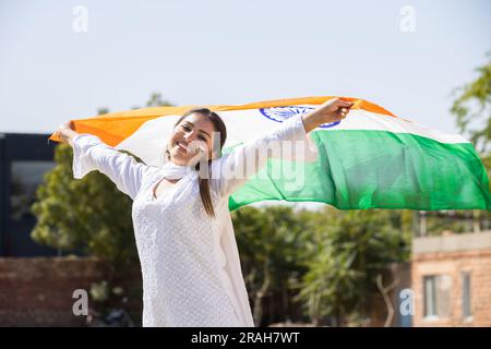 Happy young woman wearing traditional white dress weaving indian flag outdoor at park, celebrating Independence day or Republic day. Stock Photo