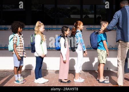 Diverse male teacher and elementary schoolchildren with schoolbags queuing in school playground. Education, inclusivity, elementary school and learnin Stock Photo