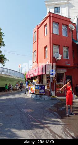 Woman in red stands beside a red building housing a kebab and ice cream shop, with people drinking Turkish tea underneath a road, Istanbul, Turkey Stock Photo