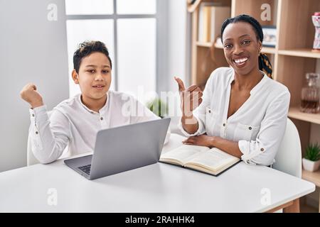 Young mother and son doing homework with laptop pointing thumb up to the side smiling happy with open mouth Stock Photo