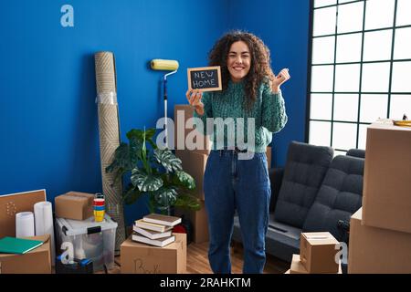 Young hispanic woman holding blackboard with new home text screaming proud, celebrating victory and success very excited with raised arm Stock Photo