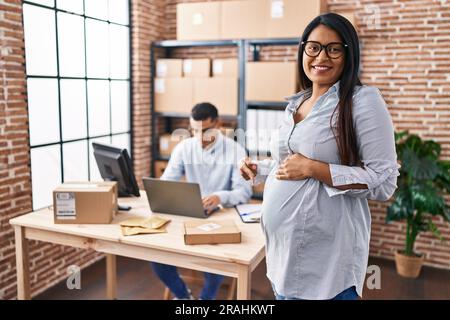 Young hispanic woman expecting a baby working at small business ecommerce smiling happy pointing with hand and finger Stock Photo