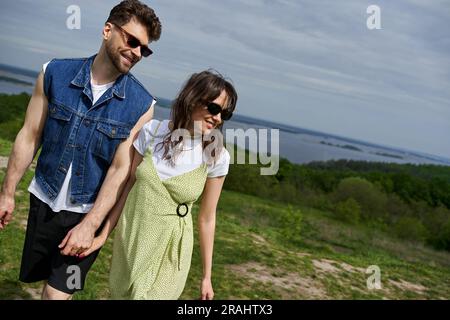Positive romantic couple in sunglasses and trendy summer outfits holding hands and looking away while walking on blurred landscape, countryside wander Stock Photo