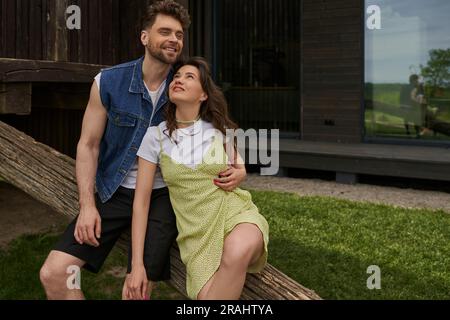 Positive an bearded man in denim vest hugging stylish girlfriend in sundress and sitting together on wooden log while relaxing in rural setting, count Stock Photo