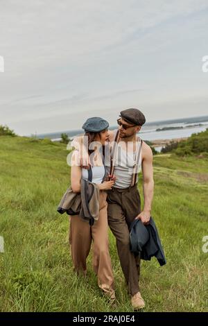 Fashionable man in vintage outfit and sunglasses hugging brunette girlfriend and talking while walking on blurred grassy hill with cloudy sky at backg Stock Photo