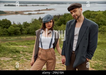Fashionable and bearded man in newsboy cap and jacket holding hand of ...