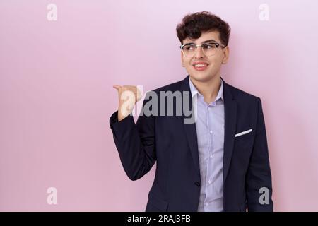 Young non binary man with beard wearing suit and tie smiling with happy face looking and pointing to the side with thumb up. Stock Photo