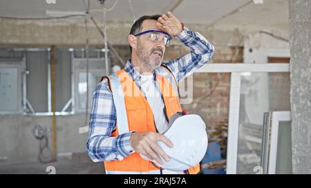 Middle age man builder holding hardhat sweating at construction site Stock Photo