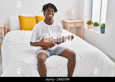 African american man playing ukelele sitting on bed at bedroom Stock Photo