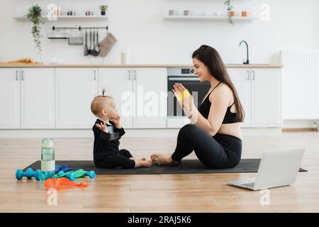 Beautiful lady and adorable child sitting with soles of feet together on floor while having fun with stretch band. Attentive baby following joyful mom's instructions during physical exercises at home. Stock Photo