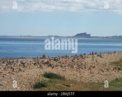 many small constructed stacks of piled stones on shore of Lindisfarne Holy Island overlooking Bamburgh Castle in distance - Northumberland, England,UK Stock Photo