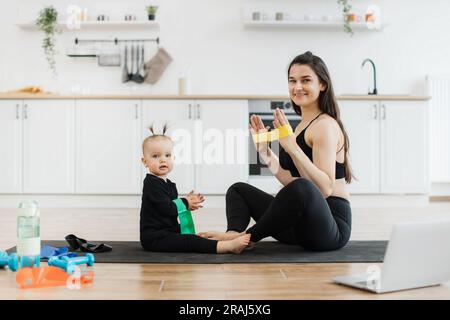 Beautiful lady and adorable child sitting with soles of feet together on floor while having fun with stretch band. Attentive baby following joyful mom's instructions during physical exercises at home. Stock Photo
