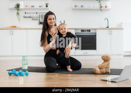 Full length view of sporty baby girl spreading legs wide apart with mom's help while smiling at camera indoors. Joyful female parent and daughter creating strong bonds while exercising together. Stock Photo