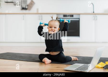 Excited infant girl in black sportswear holding dumbbells in hands