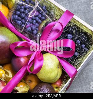 Juicy ripe fruits and berries are beautifully stacked in wooden box tied with pink ribbon. Dark background. Food delivery, gift fruit set. Concept of Stock Photo