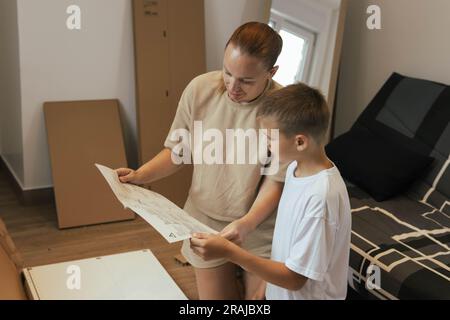 mother and child are sitting on the floor in the living room and watching the instructions to fold the closet for things.People are dressed in casual Stock Photo