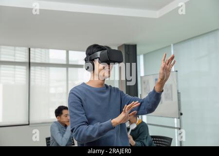 A cheerful and excited young Asian male developer tested a new VR game on a VR headset in the meeting with team. Virtual reality Stock Photo