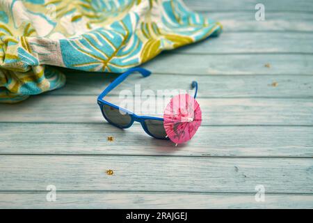 Blue sunglasses with a small pink umbrella and a tropical print towel on a wooden table. Stock Photo