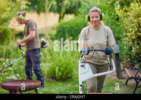 Young Female gardener using a lawn mower while her colleague or bosses is trimming the garden in the background Stock Photo