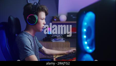 Concentrated boy in headphones plays in multiplayer game on PC at home. Gamer talks with teammates using microphone. Online video game live streaming or esports tournament. Side view. Stock Photo