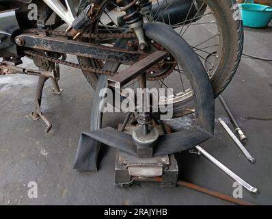 A motorcycle whose rear tire is being punctured and patched with traditional tire patching equipment in a repair shop. Stock Photo